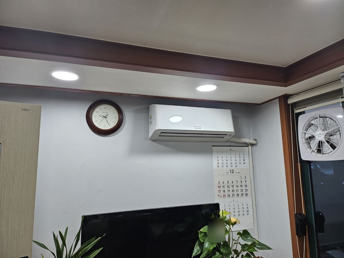 흰 벽의 둥근 시계와 벽 달력 사이 위쪽에 걸린 캐리어 벽걸이 냉난방기, 그 아래 TV와 화분